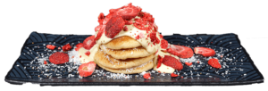 Pancakes mit weißer Schokolade, Erdbeeren und Kokosraspeln auf einem schwarzen Teller