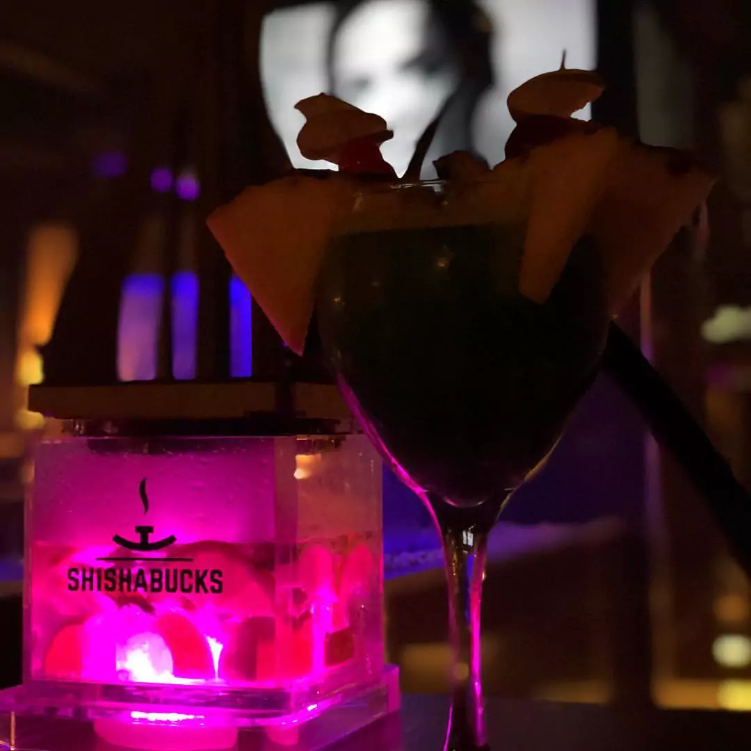 „Shisha Bar Hamburg Harburg beste“: Ein farbenfroher Cocktail, dekoriert mit Obststücken, steht neben einer beleuchteten Shishabucks-Shisha, die in magentafarbenem Licht erstrahlt. Im Hintergrund ist eine verschwommene Szene auf einem Bildschirm zu erkennen.