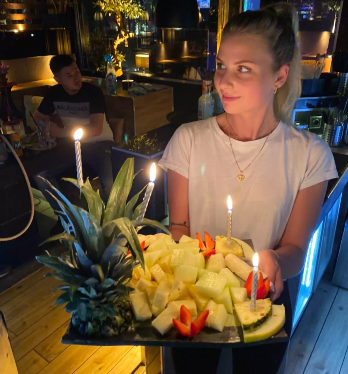 Eine Frau in einer Shisha Bar in Barmbek trägt eine dekorative Obstplatte mit brennenden Kerzen, während im Hintergrund Gäste Shisha rauchen und die entspannte Atmosphäre genießen.