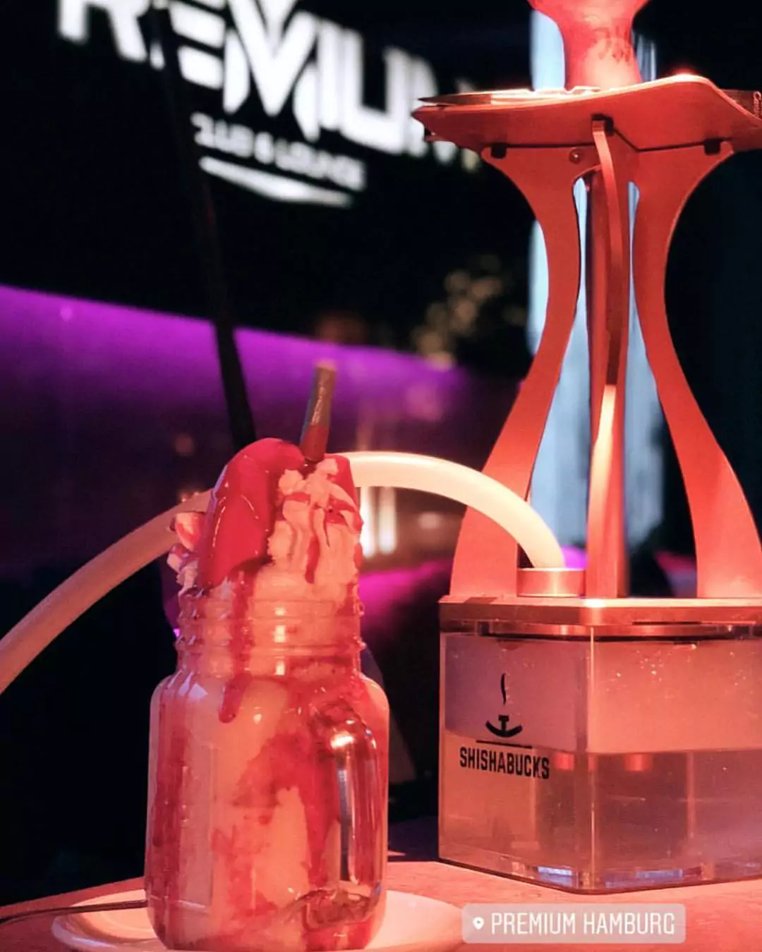 „Shisha-Bar Harburg“: Ein stilvoll angerichteter Milchshake in einem Glas mit Sahne und Erdbeersauce steht neben einer modernen Shisha von Shishabucks in einer gemütlichen, dunklen Lounge-Atmosphäre. Im Hintergrund ist das Premium Hamburg Logo sichtbar.