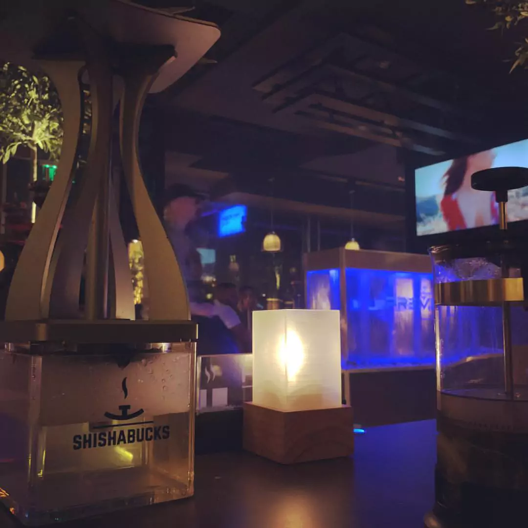 „Shisha Cafe Harburg“: Ein stilvolles Shisha-Setup mit einer Shisha von Shishabucks, einer kleinen leuchtenden Tischlampe und einem belebten Café im Hintergrund, das in gedämpften blauen und gelben Lichtern beleuchtet ist.