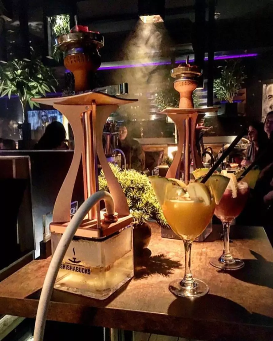 Zwei moderne Shishas und farbenfrohe Cocktails in einer gemütlichen Shisha-Bar, umgeben von sanfter Beleuchtung und Pflanzen.