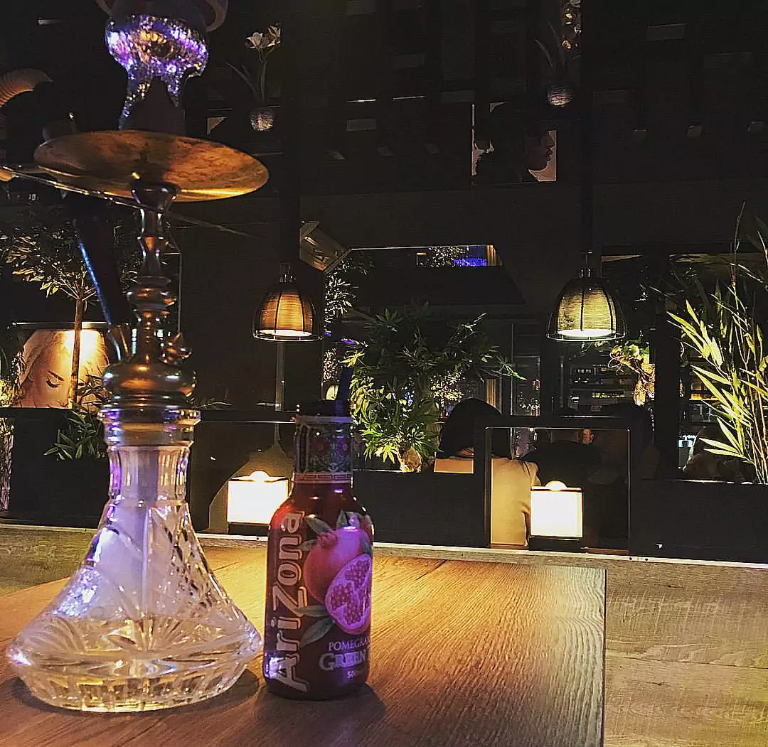 Eine Shisha und eine Arizona-Granatapfel-Getränkeflasche auf einem Tisch in der Jungfernstieg Shisha Bar, umgeben von Pflanzen und gedämpfter Beleuchtung.