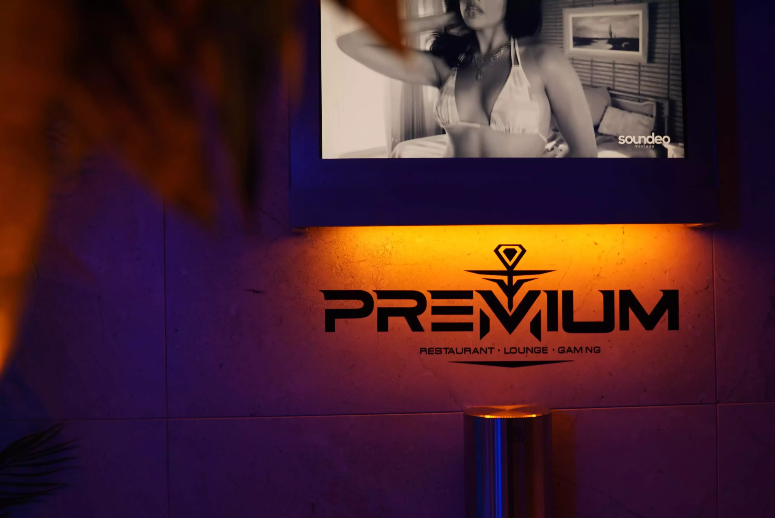 Das Premium-Logo einer Shisha-Bar, Lounge, Restaurant und Gaming-Bereich, beleuchtet von warmem gelben Licht mit einem Bildschirm im Hintergrund.